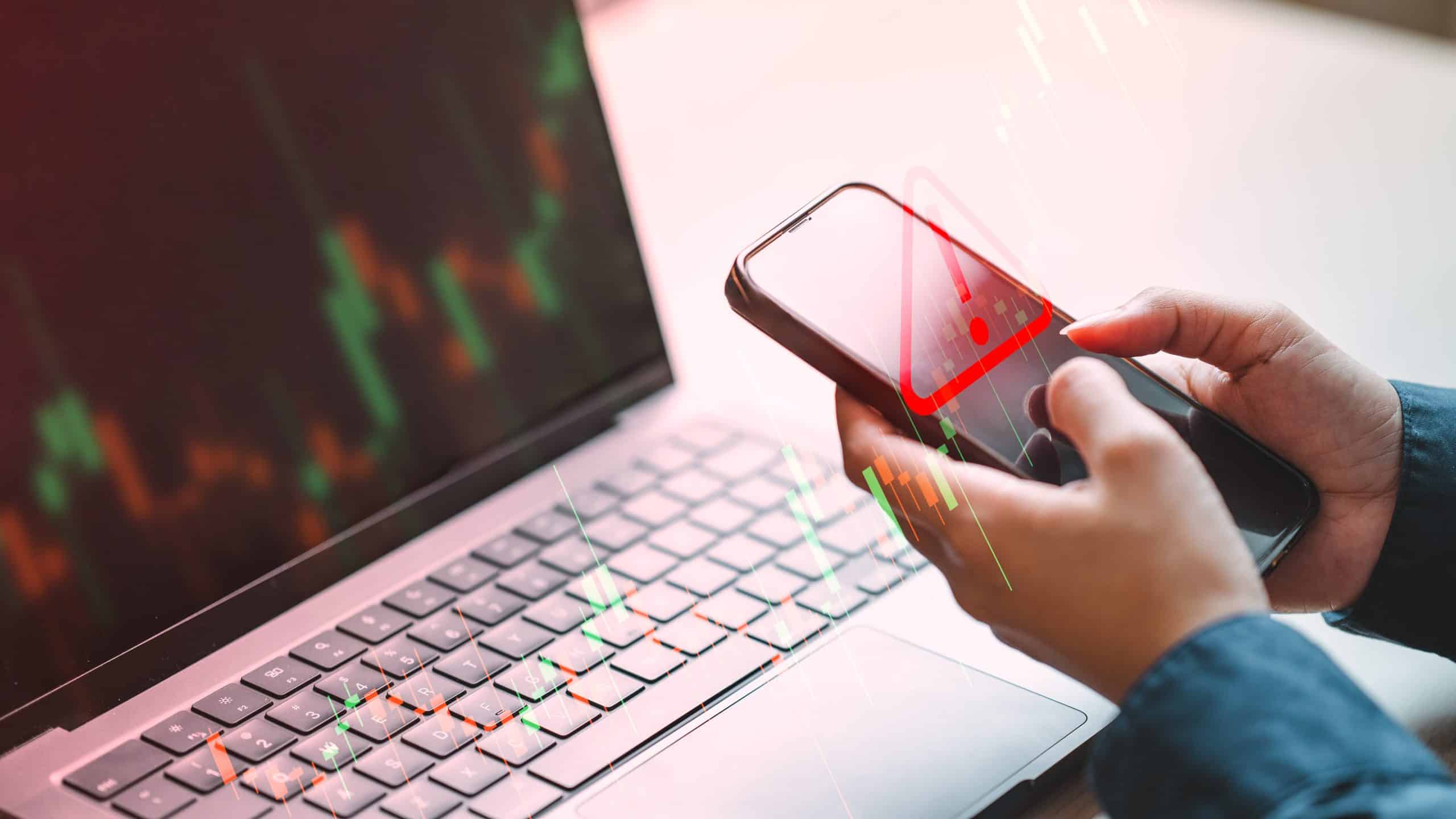 A person checks a mobile phone showing a market warning alert with falling stock charts on a laptop screen, representing risk, volatility, and sudden market downturn in financial trading.